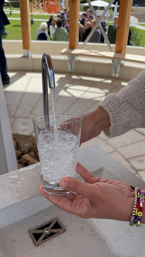 Een glas wordt gevuld met bruisend water uit een kraan; op de achtergrond zijn mensen en een paviljoen te zien.