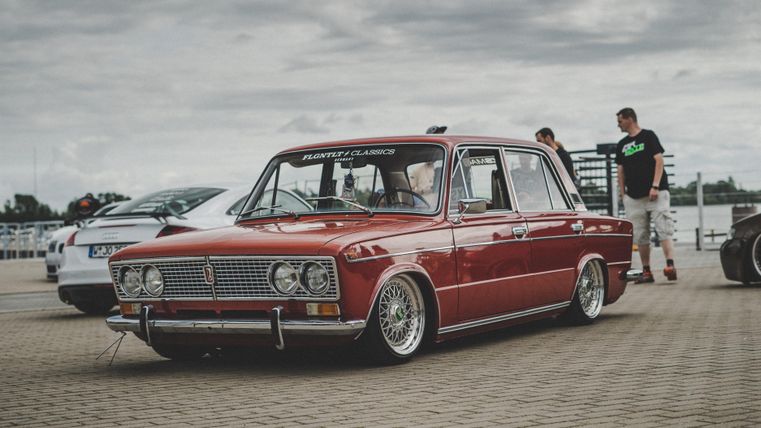 Ein roter Oldtimer steht auf einem Parkplatz. Im Hintergrund sind weitere Autos und Personen zu sehen.