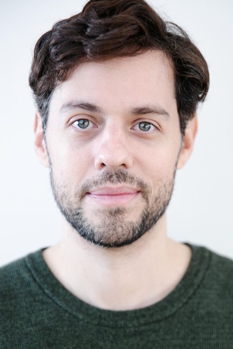 Un homme aux cheveux bouclés et à la barbe sourit à la caméra. Il porte un sweat-shirt vert et a une expression faciale amicale.