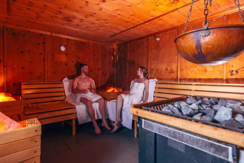 Ein Paar entspannt in einer Sauna aus Holz. Die Atmosphäre ist warm und gemütlich, mit Steinen und einem großen Topf in der Mitte.