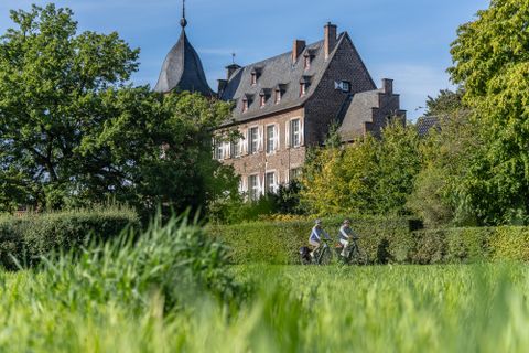 Ein malerisches Schloss umgeben von Grün und Bäumen. Radfahrer fahren auf einem Weg im Vordergrund.