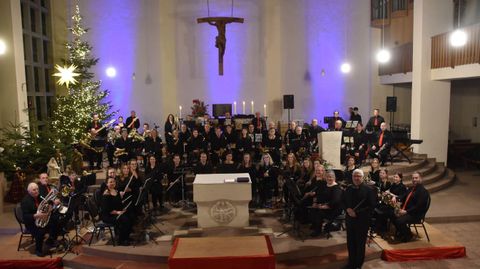 Eine große Musikgruppe steht in einer festlich geschmückten Kirche. Im Hintergrund sind ein Weihnachtsbaum und dekorative Beleuchtung zu sehen.