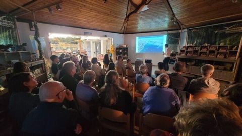 Eine Gruppe von Menschen sitzt in einem Seminarraum und hört einer Präsentation zu. Im Hintergrund ist ein Bildschirm mit Bildern sichtbar.