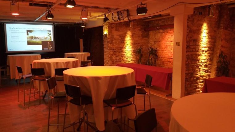 Een moderne evenementenruimte met ronde statafels en warme verlichting. Op de achtergrond zijn een projectiescherm en enkele planten te zien.