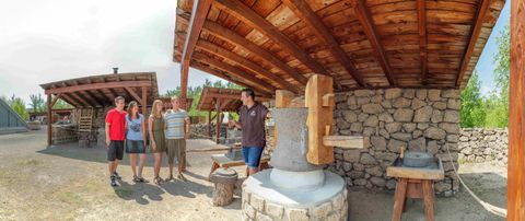 Eine Gruppe von fünf Personen steht vor einer traditionellen Mühle in einer ländlichen Umgebung. Im Hintergrund sind Holzstrukturen und eine steinige Mühlmaschine sichtbar.