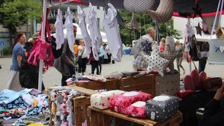 A market stall with handcrafted children's items and colorful stuffed animals. In the background, people can be seen exploring the market.