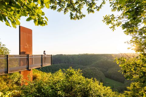 Person stands on a skywalk with a view of wooded hills at sunset.