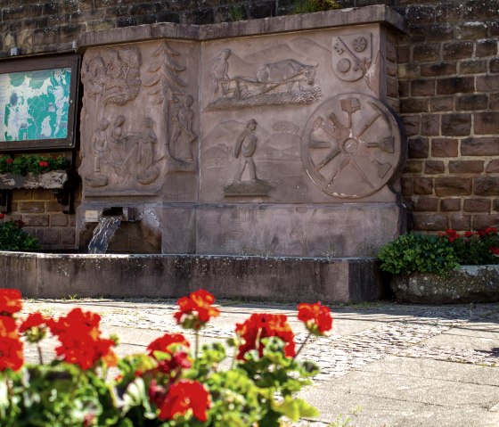 Ein Steinbrunnen mit kunstvollen Reliefs und fließendem Wasser. Rote Blumen im Vordergrund, eine Infotafel und eine Bank daneben., © TI Bitburger Land - Monika Mayer