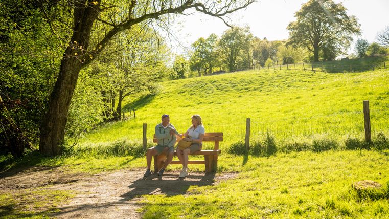 Een zonnig park met een bank waarop twee personen zitten. Op de achtergrond zijn een groene weide en bomen te zien.