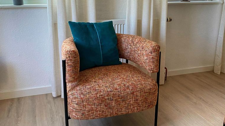 Un fauteuil moderne avec un revêtement en tissu coloré et un coussin bleu. En arrière-plan, on voit des fenêtres avec des rideaux et une petite plante.