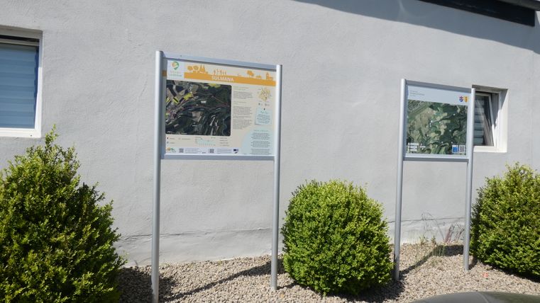 Zwei Informationstafeln an einer Hauswand mit Karten und Texten, umgeben von grünen Büschen.