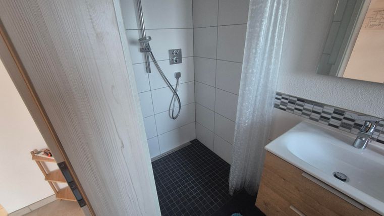 Une salle de bain moderne avec une douche et un lavabo. Le sol est en carrelage sombre et un tapis de bain bleu se trouve devant.