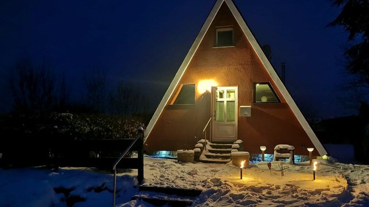 Une maison en forme de A la nuit, entourée de neige. Les lumières chaleureuses donnent à la scène une atmosphère douillette.