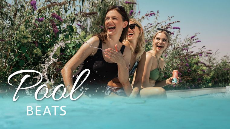 Drei fröhliche Frauen sitzen am Pool und lachen. Bunte Blumen umgeben die entspannte Atmosphäre.