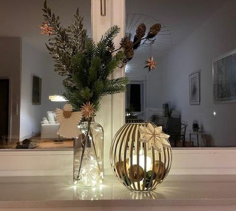 Ein festlich dekoriertes Fensterbrett mit einer Vase voller Tannenzweige und Kugeln. Eine schlichte, elegante Laterne gibt warmes Licht ab.