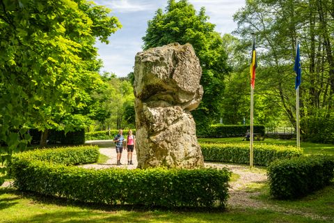 Two people are standing in front of a large rock in the green, flanked by two flags.