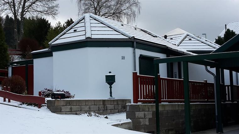 Een modern, wit huis met een plat dak, omringd door sneeuw. Op de voorgrond is een rode omheining zichtbaar die het terrein omringt.