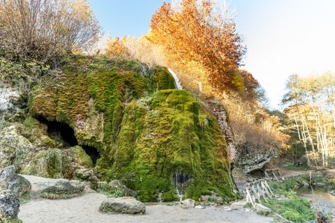 A moss-covered rock formation with gently flowing water. In the background, autumn trees shine in warm colors.