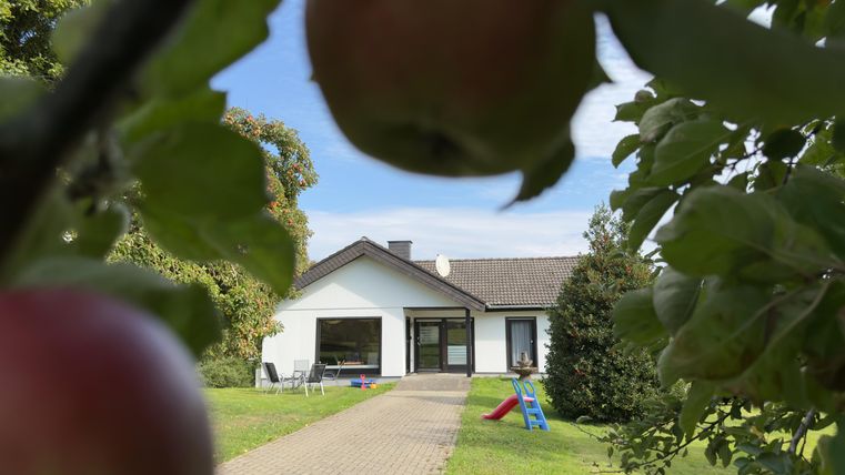 Een modern huis met een geplaveide oprit en een grote tuin. Op de voorgrond hangen appels aan een boom.