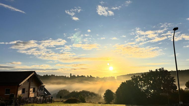 A picturesque sunrise over a misty landscape. In the foreground stands a building, surrounded by trees and gentle hills.