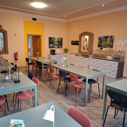 A cozy dining room with several tables and colorful chairs. In the background, there is a small kitchenette and some decorative pictures on the wall.