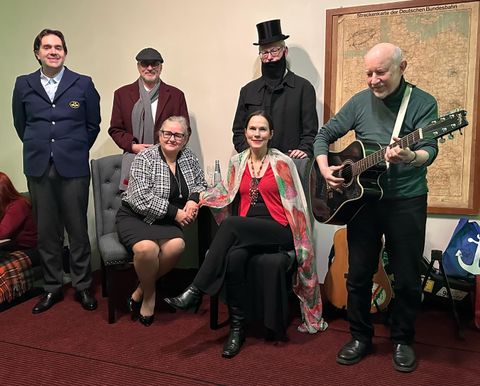 Zes personen verzamelen zich in een kamer. Drie zitten terwijl de andere drie staan, en één speelt gitaar.
