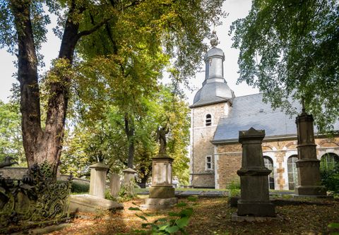 Ein Friedhof mit alten Grabsteinen und einer Kirche im Hintergrund. Die Umgebung ist von Bäumen umgeben und vermittelt eine ruhige Atmosphäre.