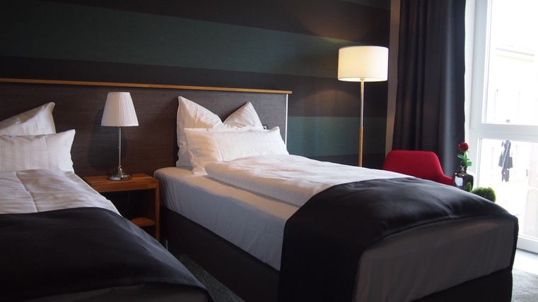 Ein modernes Hotelzimmer mit zwei Einzelbetten und einem großen Fenster. Der Raum ist mit einer Stehlampe und einem kleinen Tisch dekoriert.