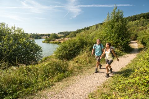 Two hikers on the path next to a lake right and left of the path green bushes, bright sky