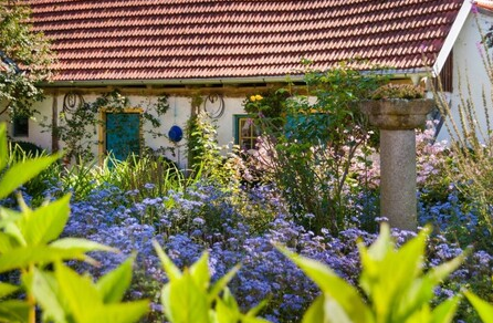 A pretty house with a red tiled roof, surrounded by colorful flowers. The bright colors and the green vegetation give the landscape an inviting atmosphere.