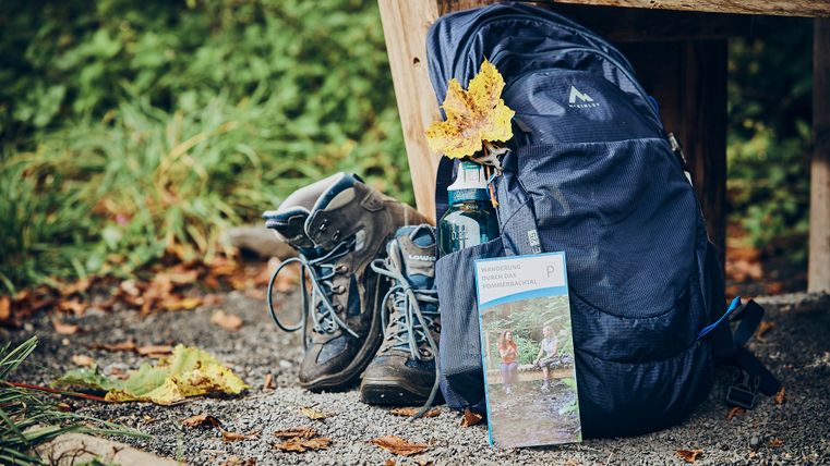 Une paire de chaussures de randonnée est appuyée contre un banc en bois, à côté d'un sac à dos bleu et d'un dépliant sur la randonnée.