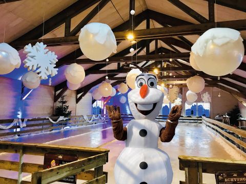 Eine Eislaufbahn mit festlicher Dekoration und einem künstlerischen Schneemann. Im Hintergrund sind große, runde Lampen und Schneeflocken zu sehen.