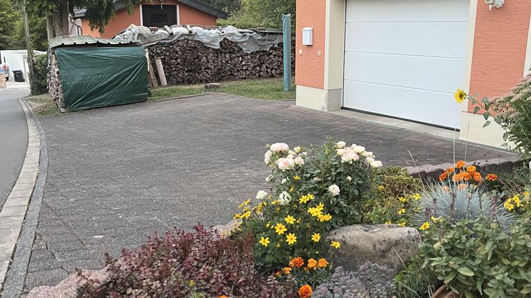 Ein gepflegter Garten mit bunten Blumen und einer runden Dekokugel. Im Hintergrund sind ein Haus und eine Garagentür sichtbar, umgeben von grünen Bäumen.
