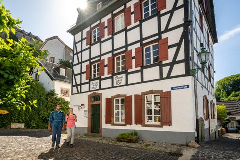 Ein charmantes Fachwerkhaus mit weißen Wänden und roten Fensterläden. Zwei Personen spazieren gemächlich die Straße entlang.