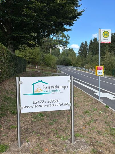 A signpost for vacation apartments called "Haus Sonnentau" stands by a road. Next to it, there is a bus stop in a rural setting.