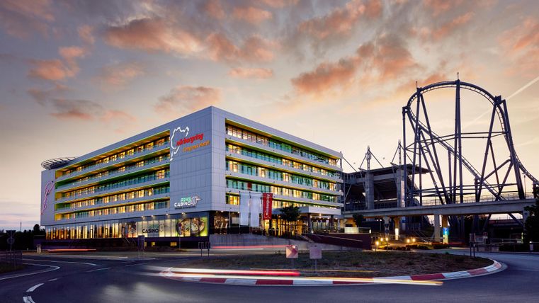 Ein modernes Hotelgebäude mit bunten Fenstern und einer großen Achterbahn daneben. Der Himmel ist abends in warmen Farben getönt.