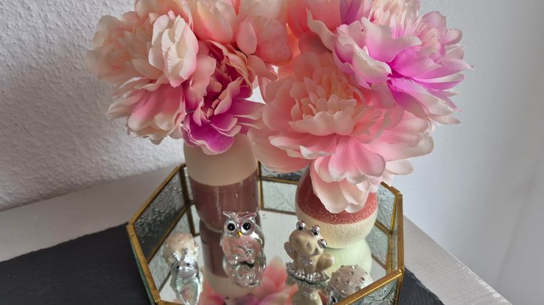 Een decoratieve container met kunstbloemen in zachte roze en crème tinten. Ook zitten er kleine figuren in de vorm van uilen en andere dieren in.
