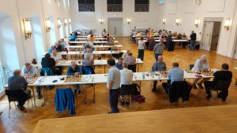 Eine Gruppe von Schachspielern sitzt an Tischen in einem großen Raum. Es herrscht eine konzentrierte Atmosphäre während des Spiels.