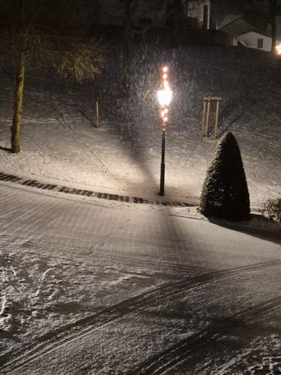 Eine schneebedeckte Landschaft bei Nacht mit einem beleuchteten Laternenpfahl. Im Hintergrund sind schneebedeckte Bäume und ein Hügel zu sehen.