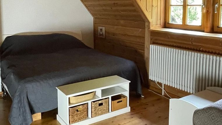 A cozy bedroom with a double bed and wood details. The room has large windows and a modern chest of drawers.