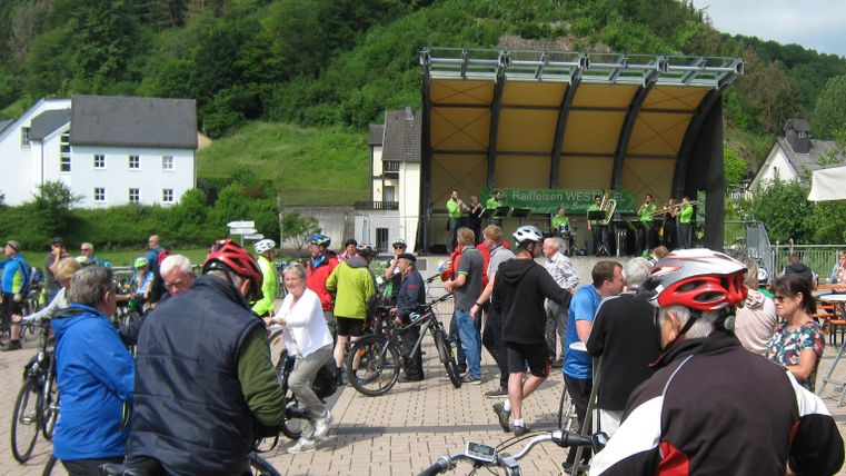 A group of cyclists gathers in a plaza in front of a stage. In the background, green hills and buildings can be seen.