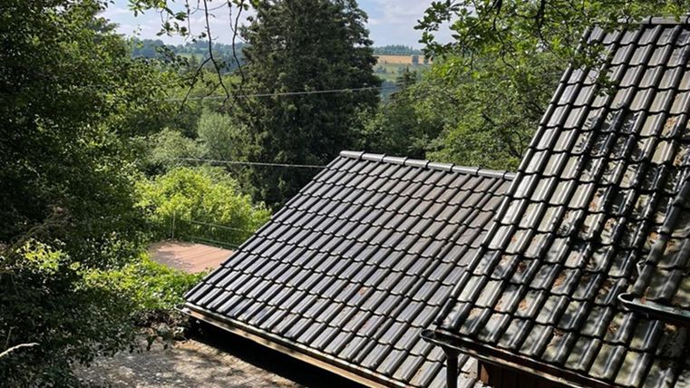 A view of a roof with slate-like tiles, surrounded by trees and greenery. In the background, gentle hills and a clear sky can be seen.