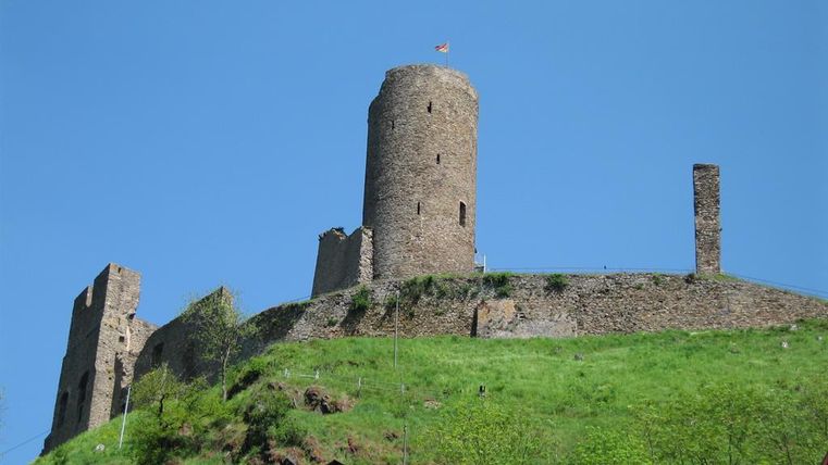 Eine alte Burg auf einem Hügel, umgeben von grüner Wiese. Der Himmel ist klar und blau.