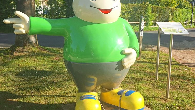 A colorful statue of a smiling character in a green shirt and a cap. The character is standing on a lawn and pointing forward with a finger.