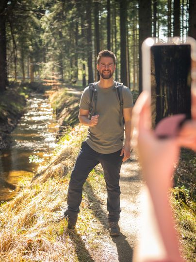 Un homme souriant se tient sur un sentier forestier à côté d'un petit ruisseau. Il tient un téléphone portable à la main et pose pour une photo.