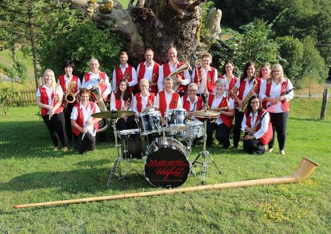 Eine Musikkapelle mit etwa 20 Mitgliedern steht im Freien, umgeben von Bäumen. Alle tragen rote Westen und präsentieren verschiedene Musikinstrumente.