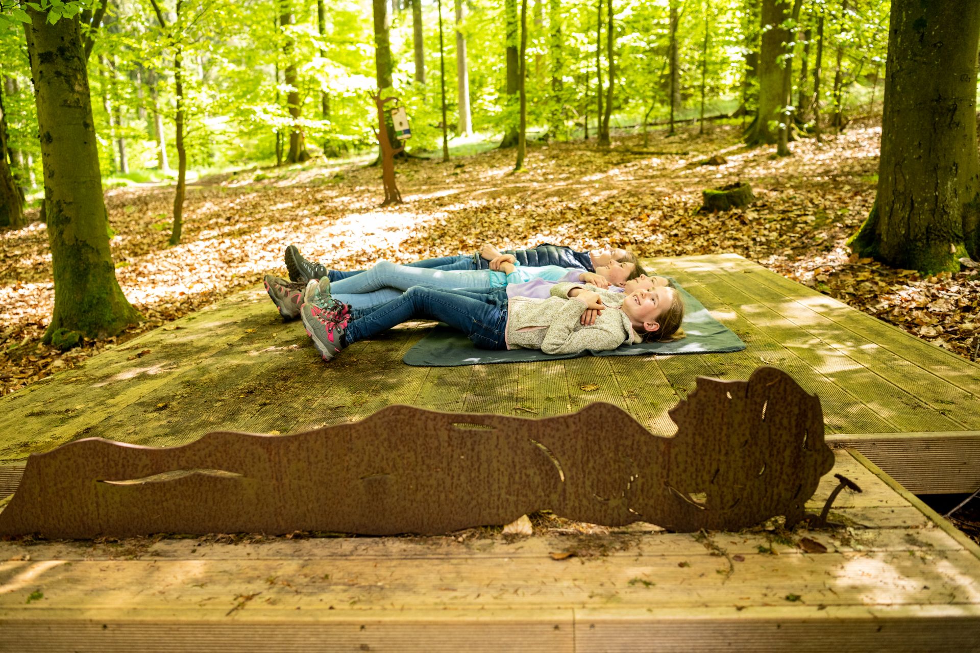 Drei Kinder liegen entspannt auf Matten auf einer Holzplattform mitten im hellen Wald und genießen eine Pause auf der Wanderung. Im Vordergrund steht ein rustikales Metall-Silhouettenobjekt in Tierform.