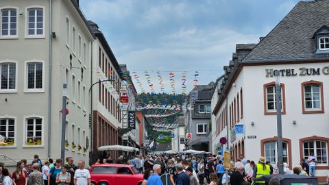 Een drukke straat met veel mensen en kleurrijke vlaggen. Aan beide zijden staan historische gebouwen en tafels van cafés zijn neergezet.