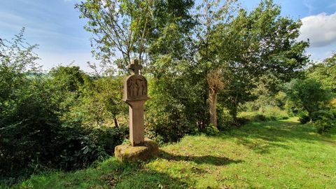 Stone cross on a meadow, surrounded by trees and bushes, under a blue sky.