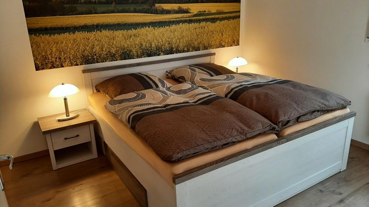 Ein gemütliches Schlafzimmer mit einem Doppelbett und sanften Lichtern. Im Hintergrund ist ein großes Landschaftsbild mit Feldern zu sehen.